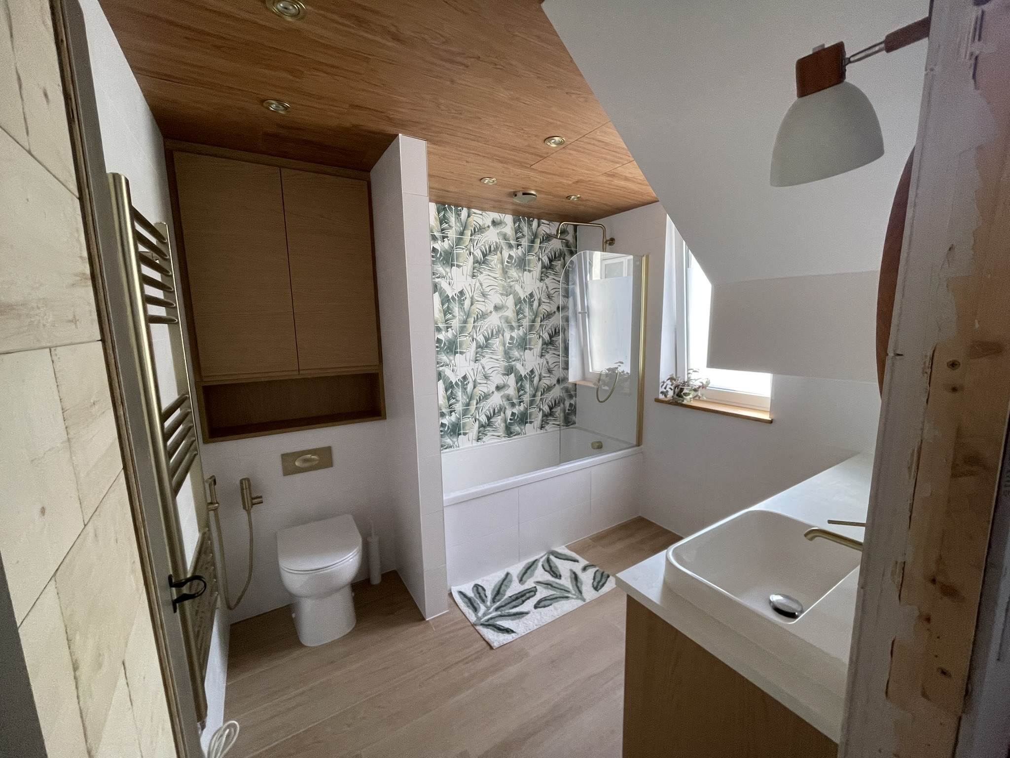 Loft bathroom with oak vanity unit, botanical print tiles, gold fixtures and bath by KDB Home Solutions