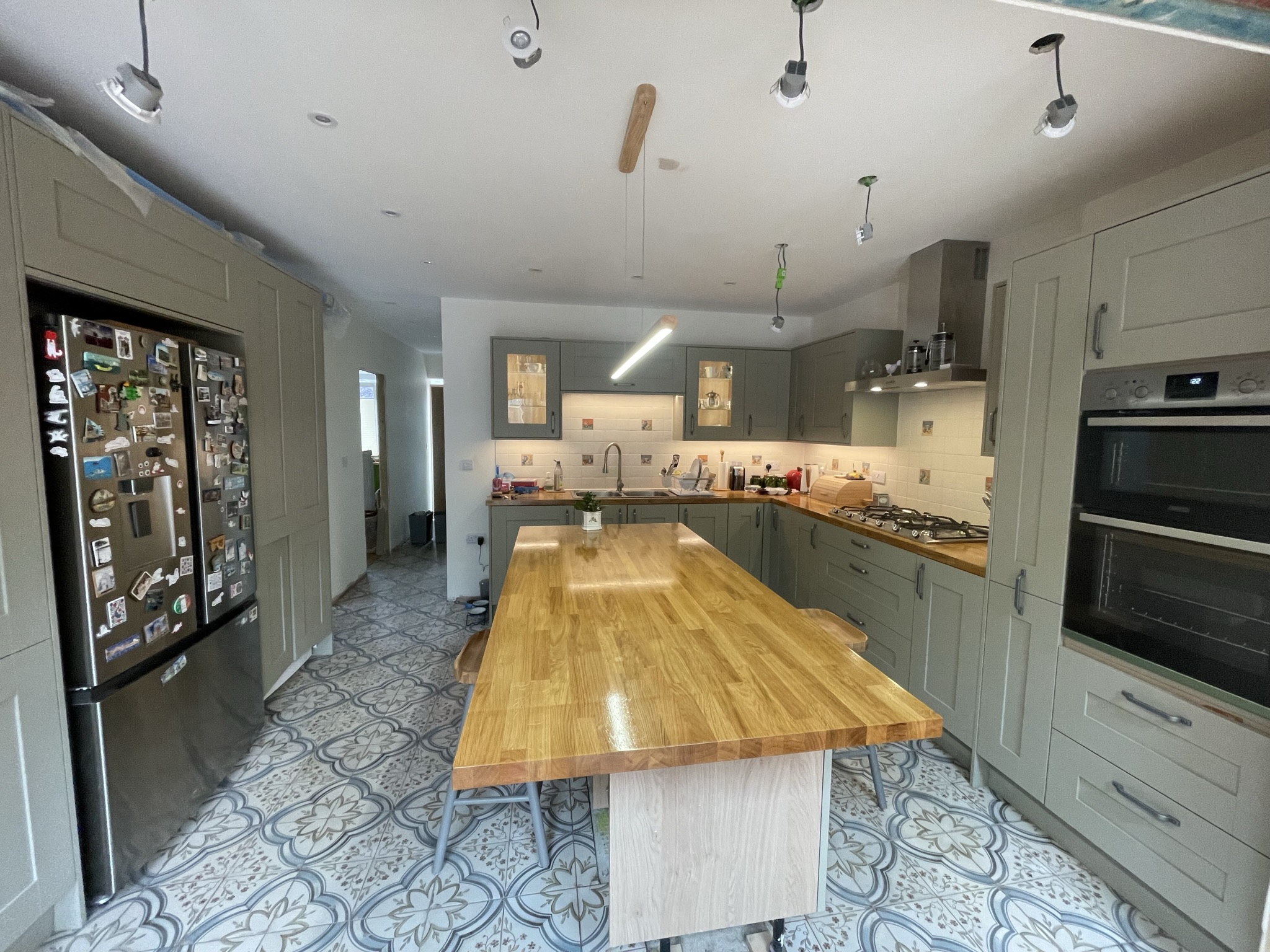 Large grey-green shaker kitchen with oak dining island, double oven and ornate patterned floor tiles by KDB Home Solutions