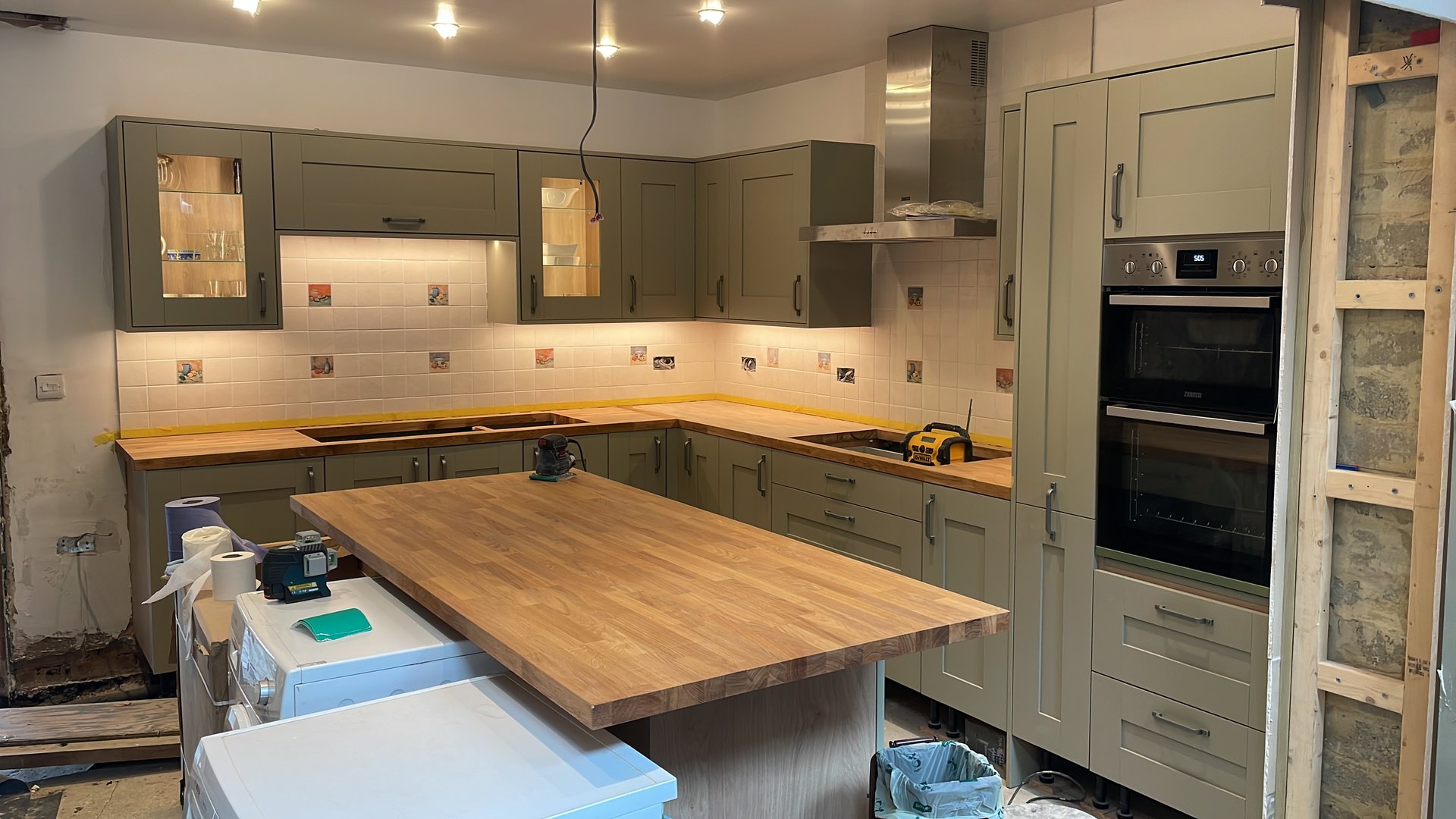 Sage green shaker kitchen during installation with central island, oak worktop and double oven by KDB Home Solutions